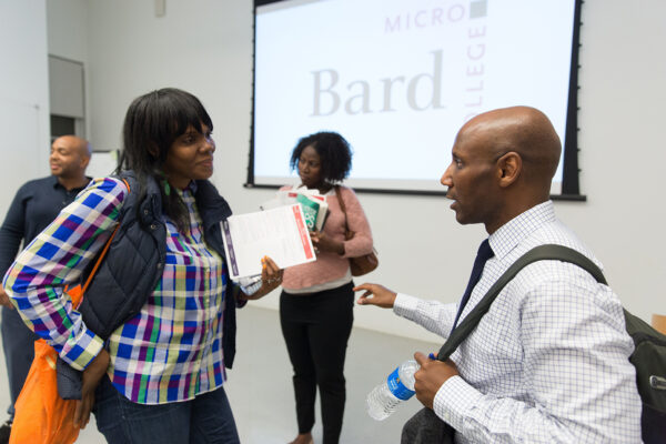 Female with papers talking to a man with a backpack on, with the Bard Microcollege logo projected onto a screen in the background.