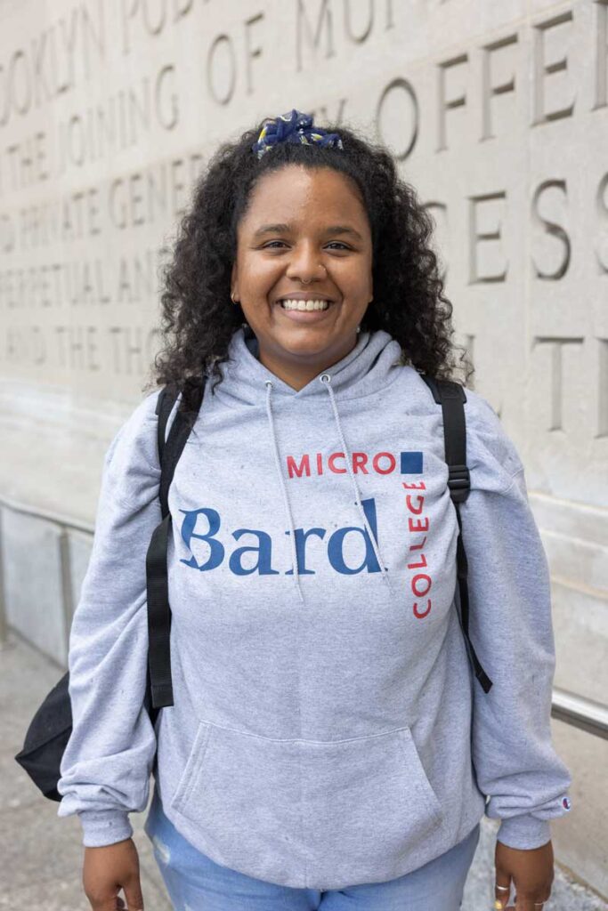 Student in microcollege sweatshirt smiling on the steps of the Brooklyn Public Library.